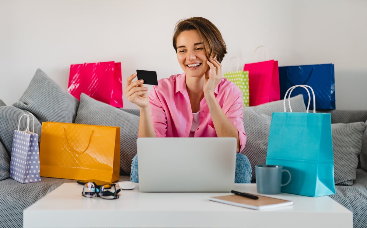 feliz-mujer-sonriente-camisa-rosa-sofa-casa-coloridas-bolsas-compra-tarjeta-credito-pagando-linea-computadora-portatil