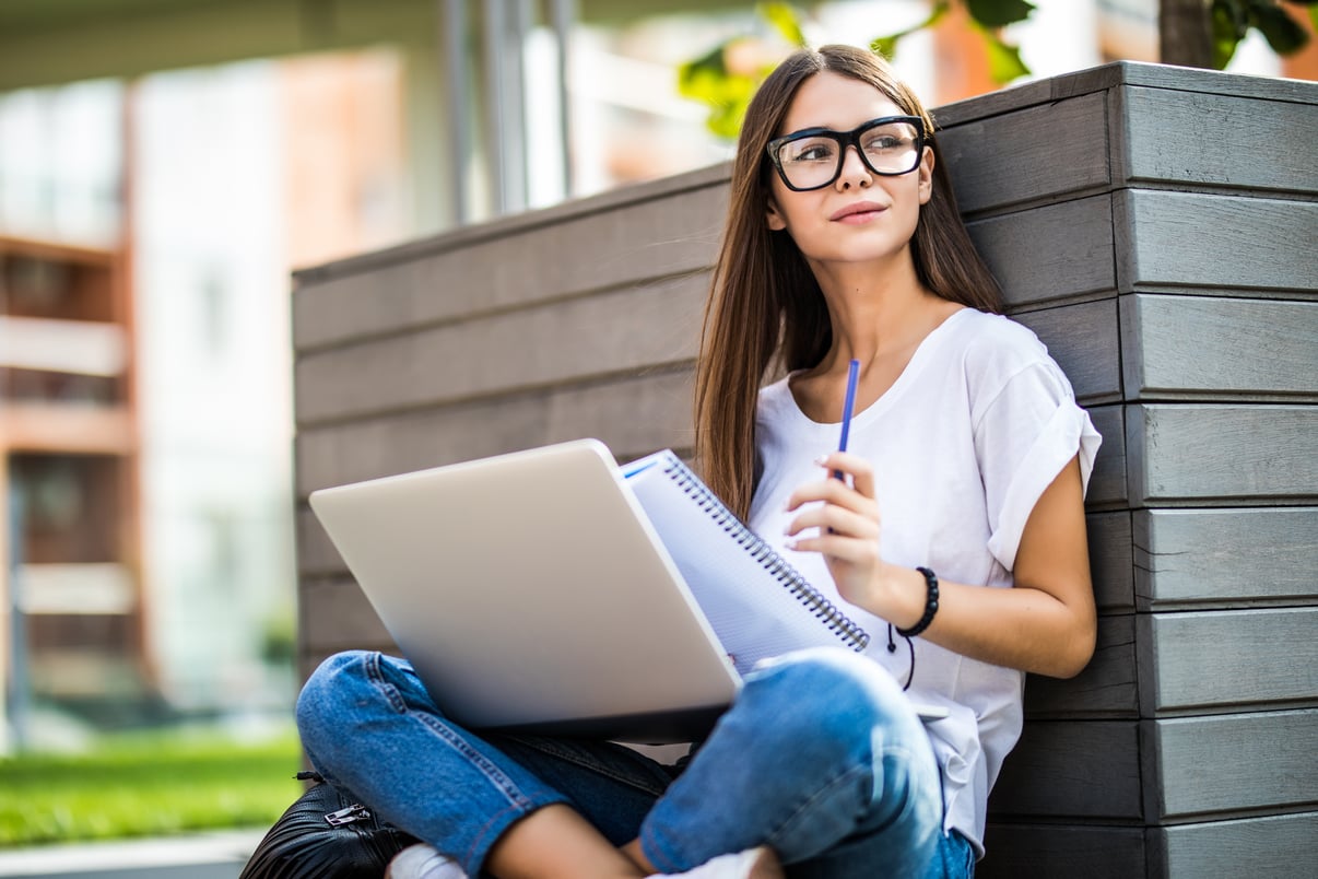 mujer-joven-feliz-ropa-casual-gafas-usando-computadora-portatil-moderna-tomando-notas-mientras-sentado-banco-calle-ciudad